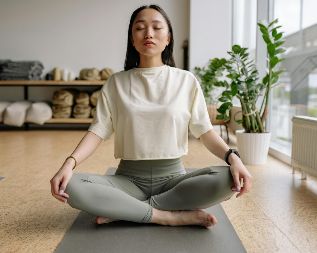Woman sitting cross-legged on a yoga mat indoors, eyes closed, conveying peace and relaxation.
